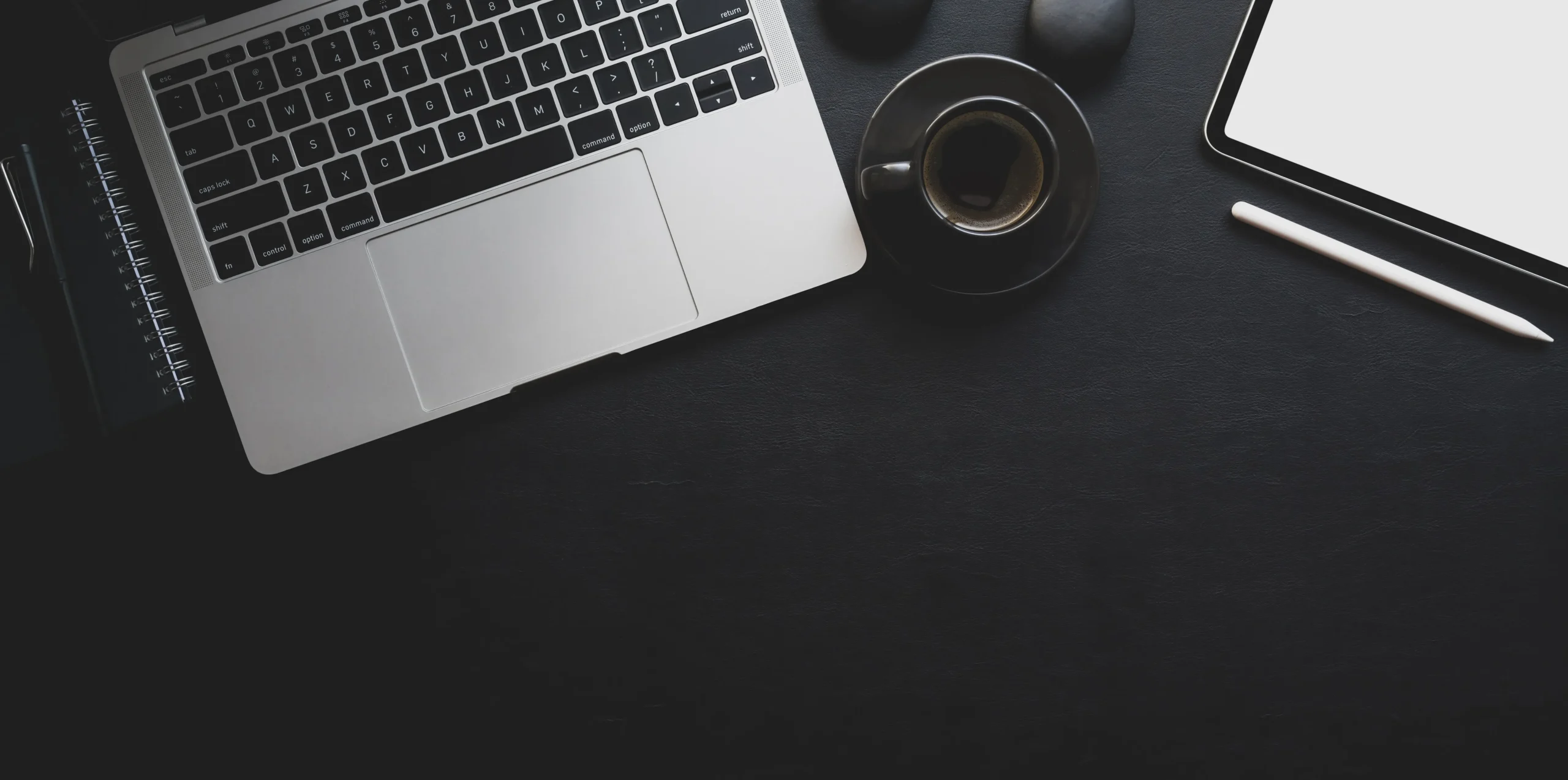 A laptop, a cup of coffee, a smartphone, and a pencil rest on a dark desk surface. The laptop is open, revealing part of the keyboard next to the blank phone screen. This modern and minimalist workspace could easily belong to an insurance professional in their element.