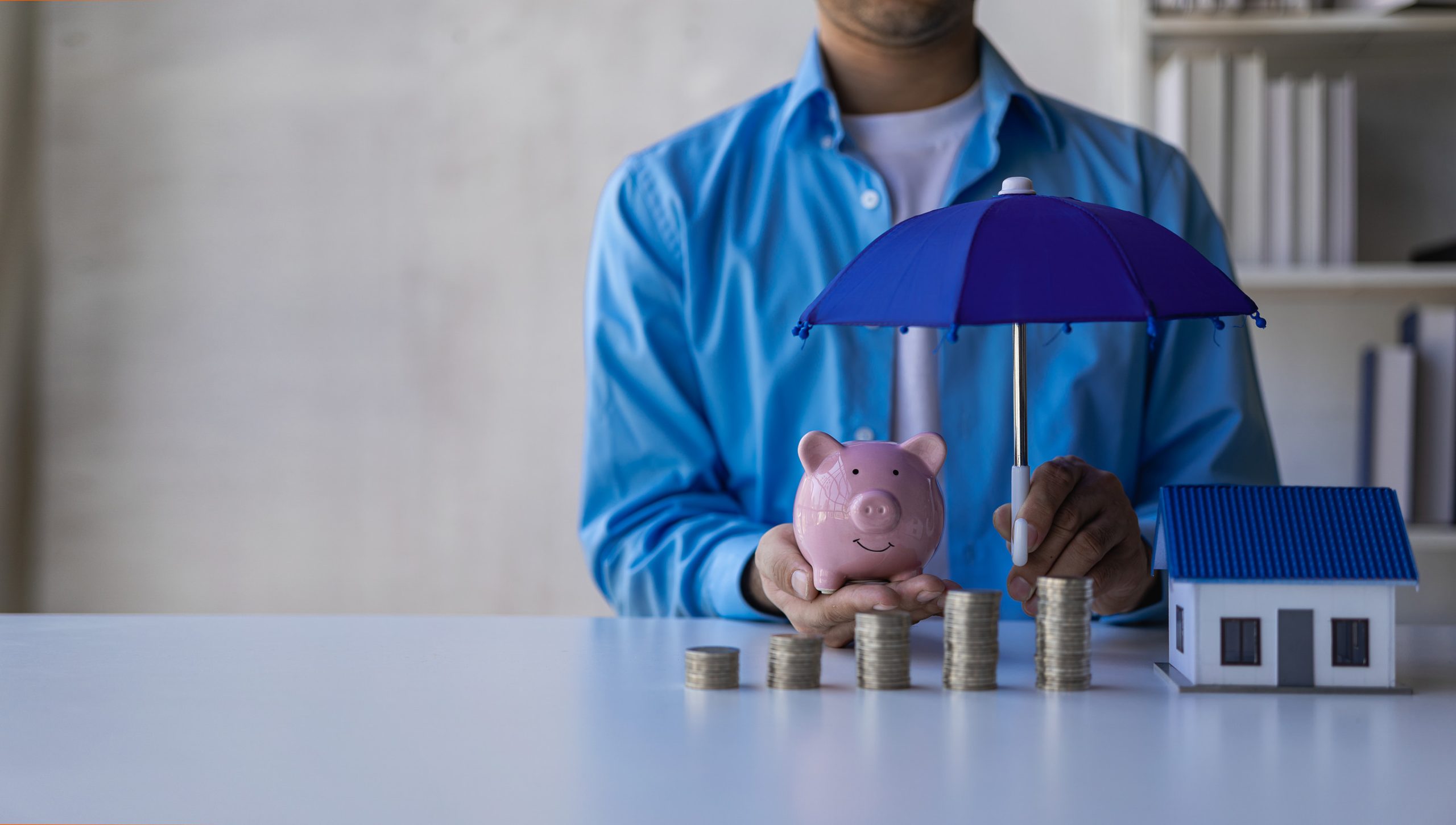 A person in a blue shirt is holding a pink piggy bank and a blue umbrella over stacked coins, with a small house model on the right. The scene suggests how affects insurance by emphasizing financial savings and protection.
